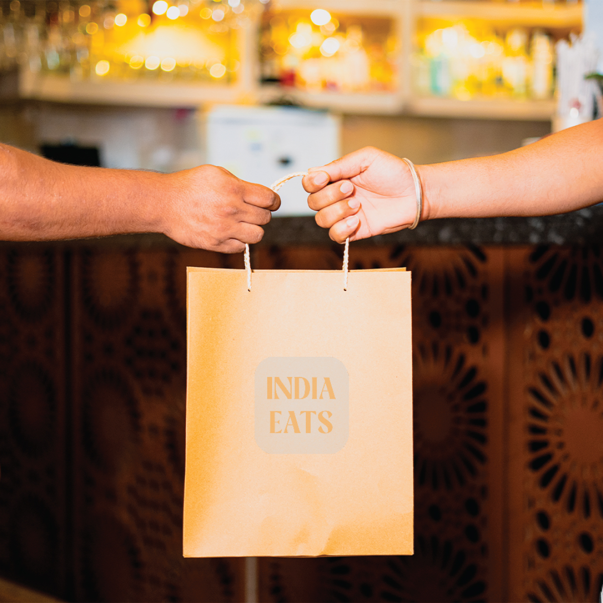 Two hands exchanging orange paper bag labeled "India Eats".
